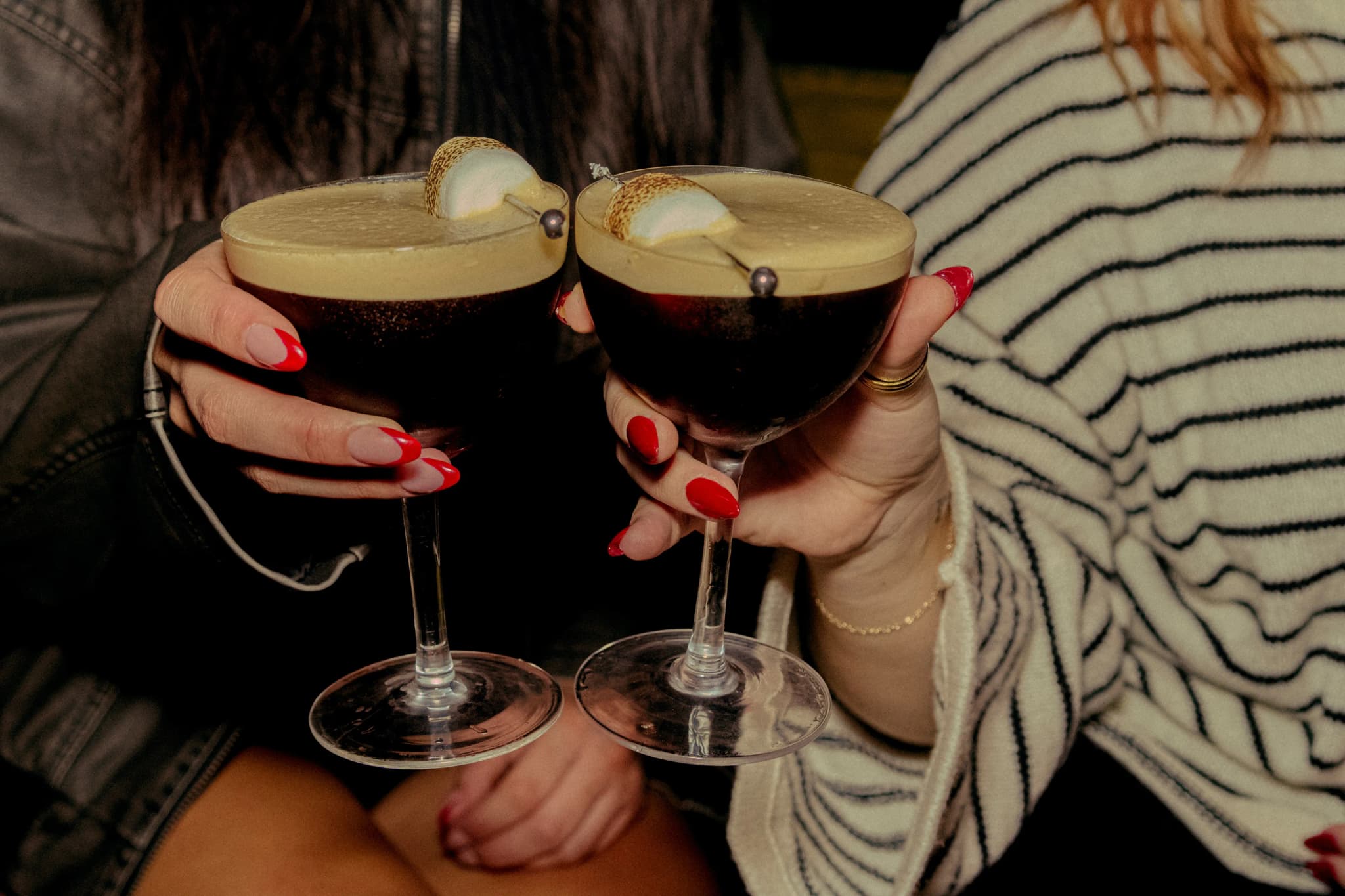 Twee jongvolwassenen proosten met koffiecocktails in een gezellige binnenomgeving, elk met een marshmallow en parelbolletje bovenop.