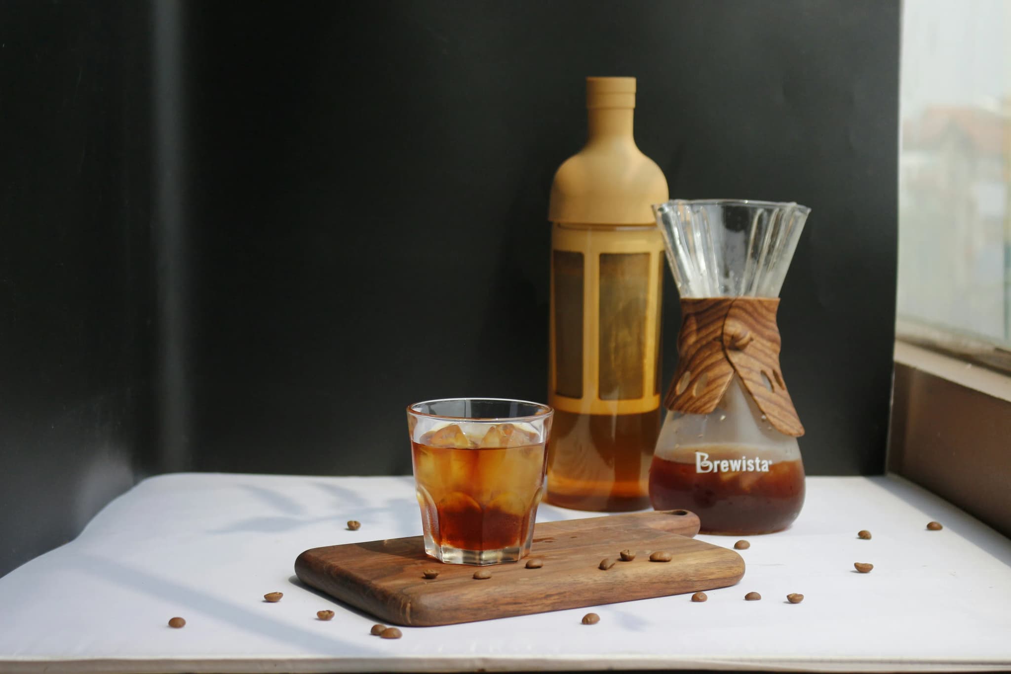 Close-up van een glas cold brew koffie met ijs op een houten plank, omringd door koffiebonen en koffie-accessoires op een donkere achtergrond.