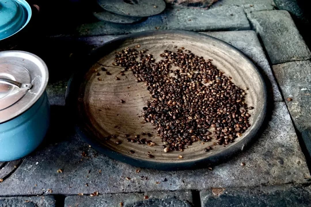 Geroosterde koffiebonen op een ronde schaal met slijtsporen, omgeven door oude kookpotten op een ruwe stenen ondergrond.