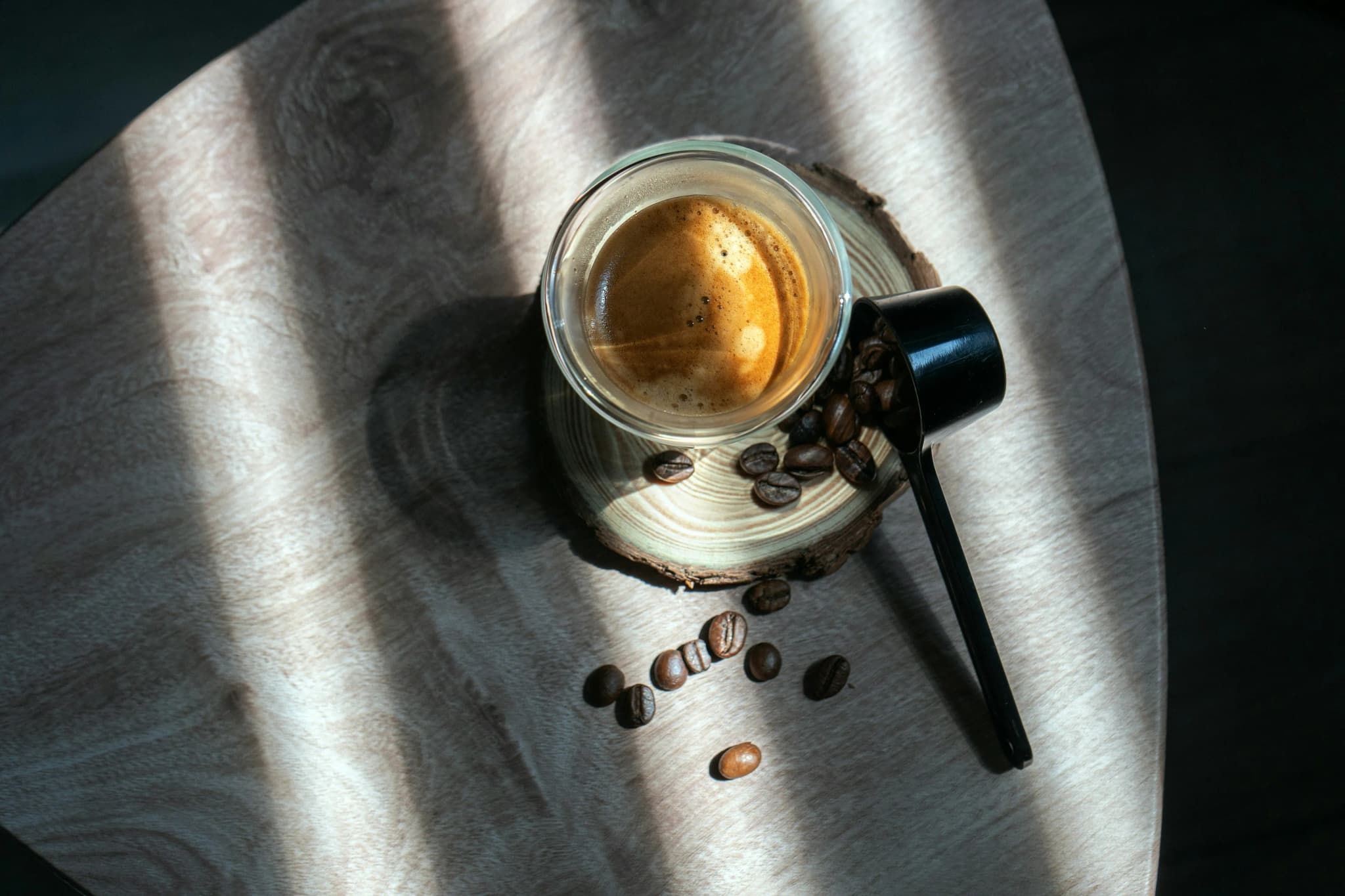 Glazen tas met espresso en crema op houten onderzetter, rondom koffiebonen en een schep, belicht op een glanzend metalen oppervlak.